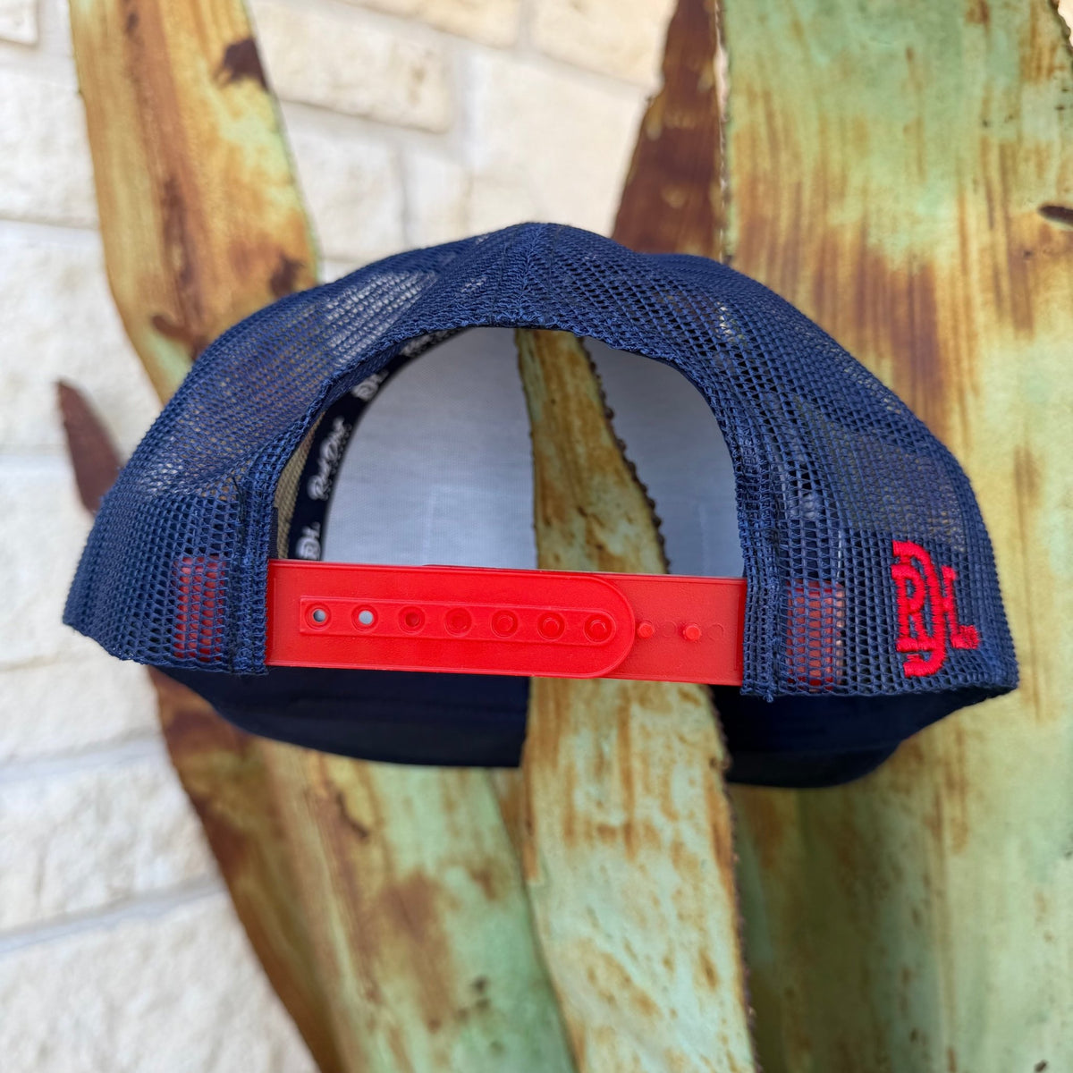This Red Dirt Hat Co. trucker keeps it classic with a structured navy front, breathable mesh back, and a bold embroidered patch that does all the talking. The red rope accent cord along the brim adds just enough detail to set it apart from every other cap on the rack.