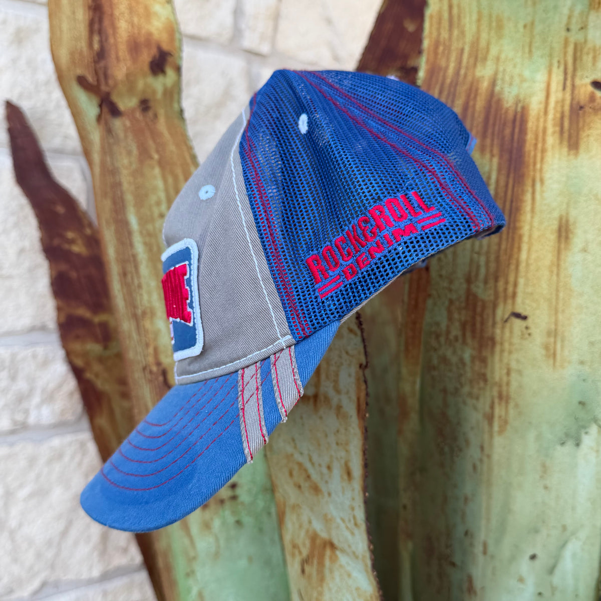 A blue and gray baseball cap with red stitching and 'ROCK & ROLL DENIM' embroidered on the side, leaning against rustic wooden branches.