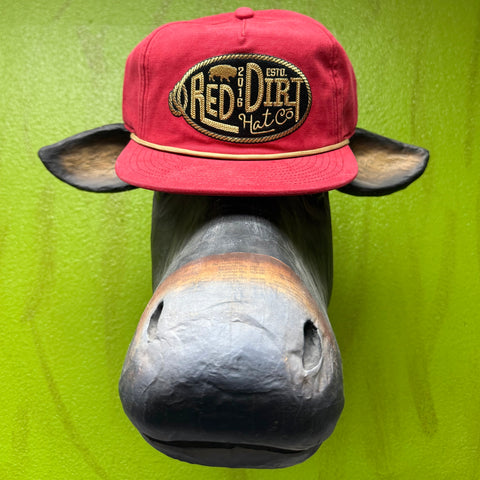 Bold logo, clean lines, Western attitude. This Red Dirt Hat Co. snapback features a structured red canvas crown and a standout oval embroidered patch framed in gold rope — the kind of cap that looks sharp whether you're at the ranch, the rodeo, or the restaurant. The gold rope accent cord along the brim ties it all together without trying too hard.