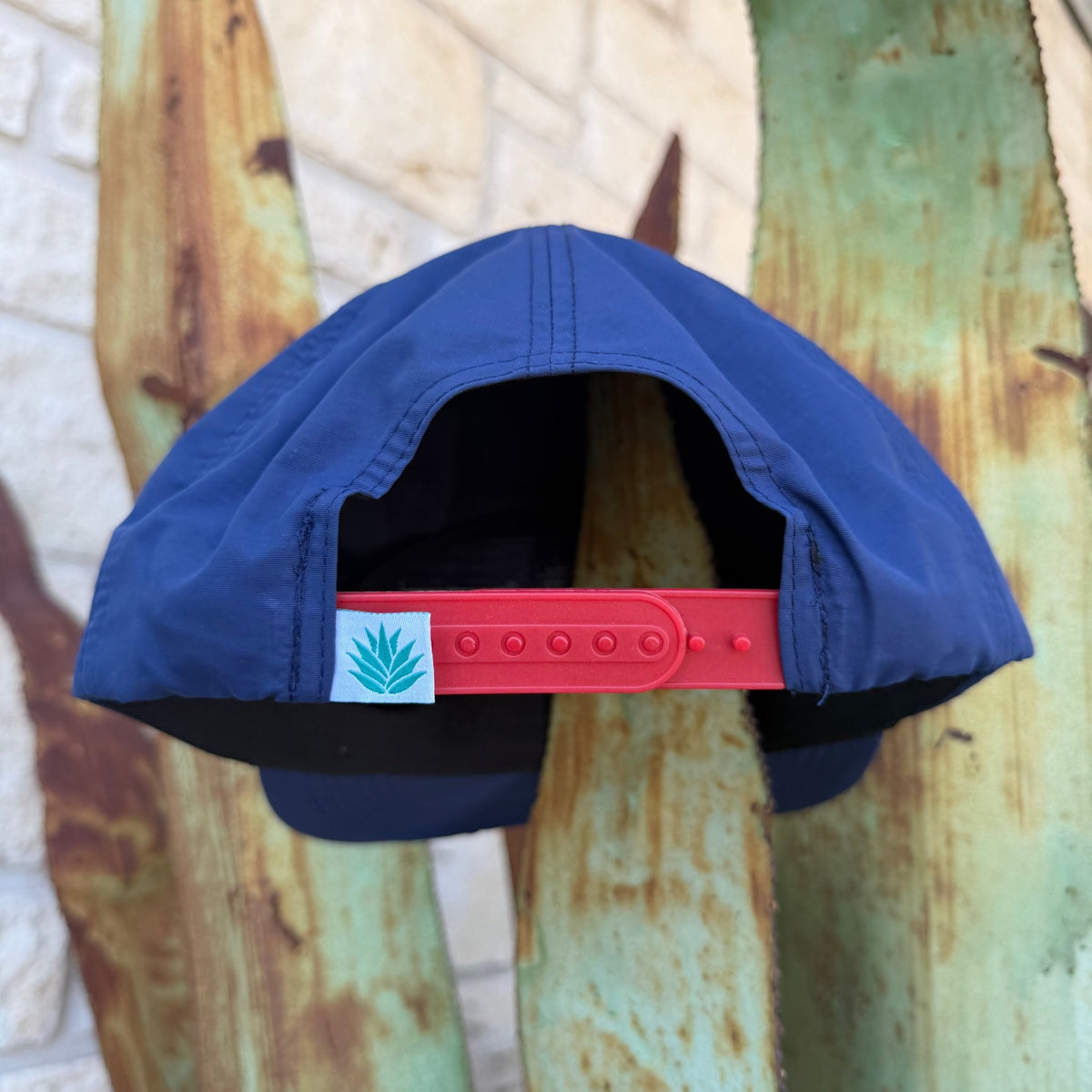 A blue cap viewed from the back, featuring a red adjustable strap closure with a small white square patch printed with the Sendero Logo 