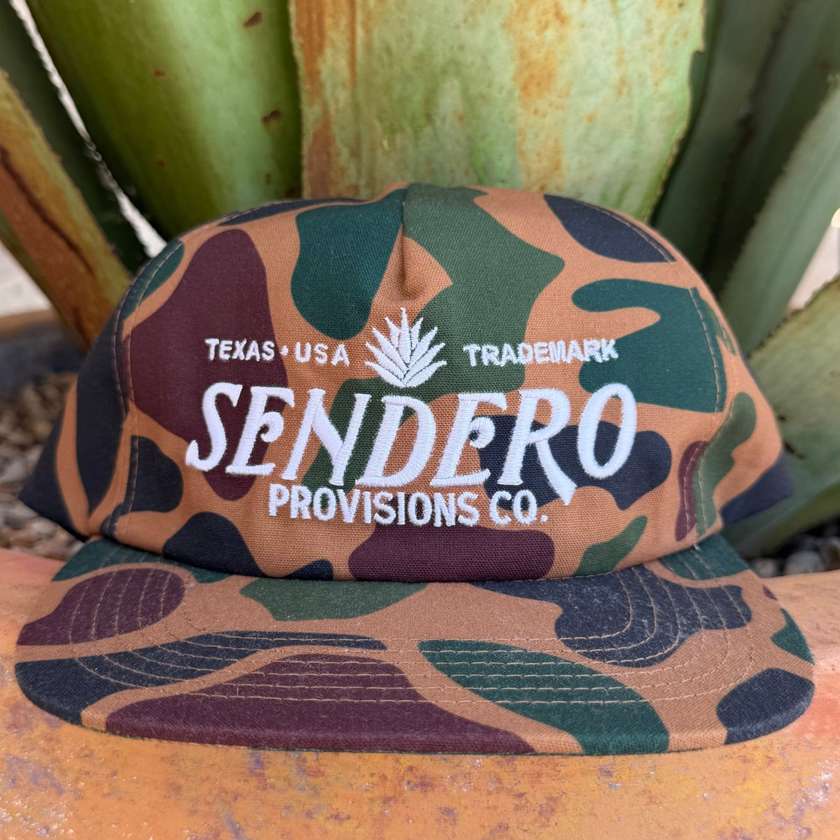 A camouflage cap with white embroidered text reading 'TEXAS USA TRADEMARK SENDERO PROVISIONS CO.' and the Sendero logo  on the front, 

