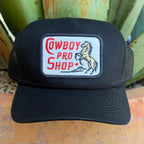 A black trucker hat with a rectangular embroidered patch on the front that reads 'Cowboy Pro Shop' in red and shows a rearing horse.