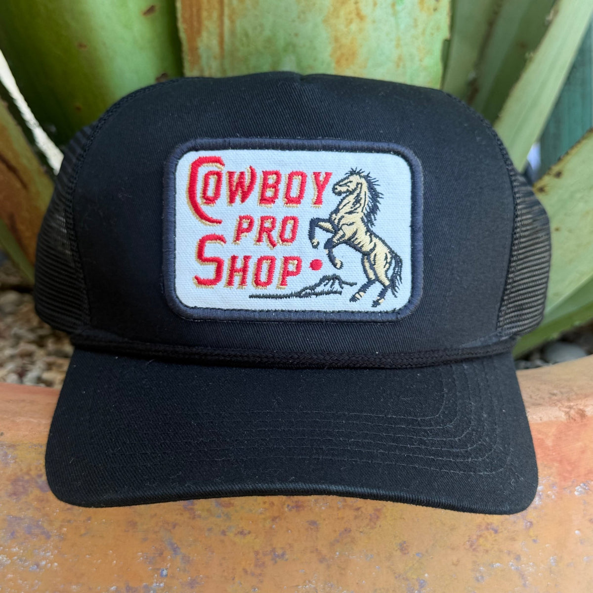 A black trucker hat with a rectangular embroidered patch on the front that reads 'Cowboy Pro Shop' in red and shows a rearing horse.