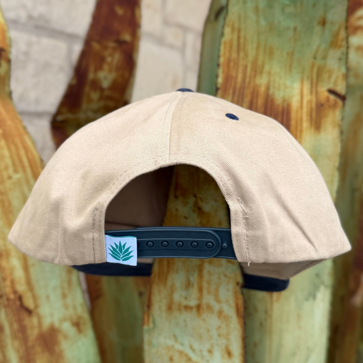 A beige snapback cap shown from the back, featuring a dark navy blue adjustable strap and a small white square patch with the Sendero logo 