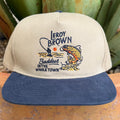 A beige and navy blue snapback cap featuring an embroidered design of a fisherman and a largemouth bass with the text 'Leroy Brown' and 'Baddest IN THE WHOLE TOWN'.