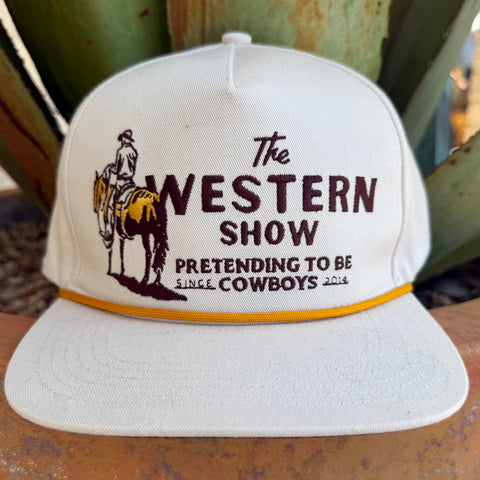 An off white snapback cap with a brown and yellow embroidered graphic reading 'The Western Show Pretending to Be Cowboys 2014' and an illustration of a cowboy on a horse,