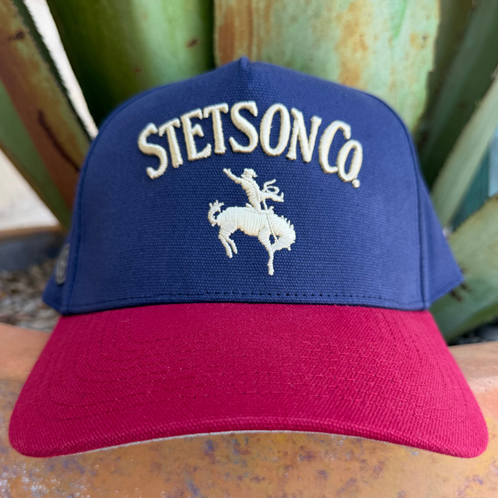 A navy blue and redr trucker hat with 'STETSON CO.' and a bucking rider embroidered on the front, displayed outdoors in front of green agaves.