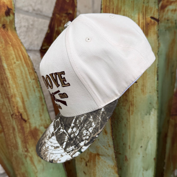 A white baseball cap with brown embroidered text and a rifle graphic, paired with a gray and black camouflage patterned visor