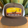 A brown and white trucker hat with a front patch reading 'RED DIRT HAT CO.' and showing a sunset landscape with a bird, clouds, and a windmill