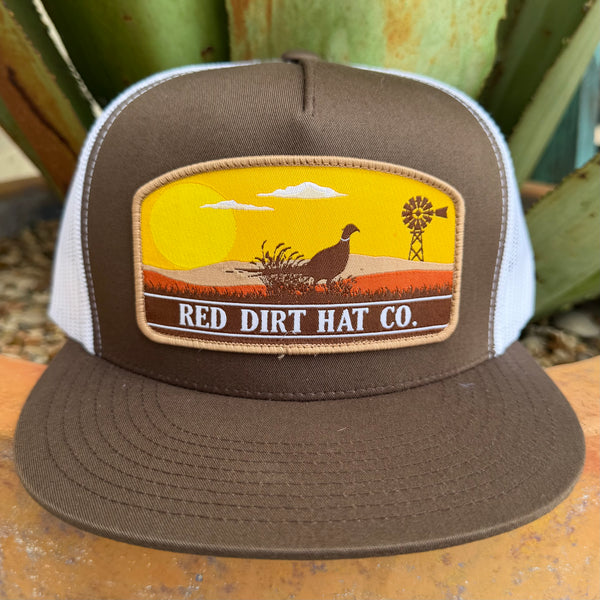 A brown and white trucker hat with a front patch reading 'RED DIRT HAT CO.' and showing a sunset landscape with a bird, clouds, and a windmill