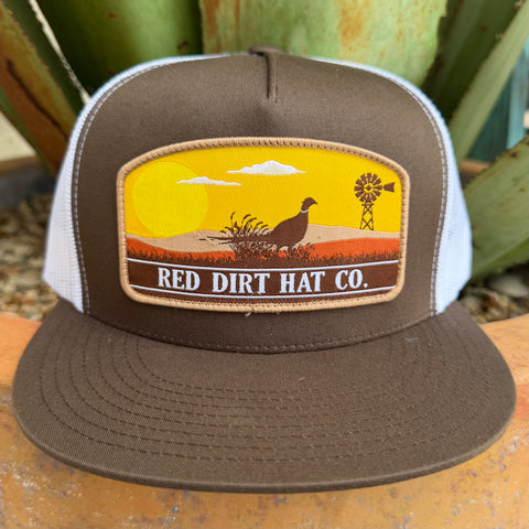 A brown and white trucker hat with a front patch reading 'RED DIRT HAT CO.' and showing a sunset landscape with a bird, clouds, and a windmill