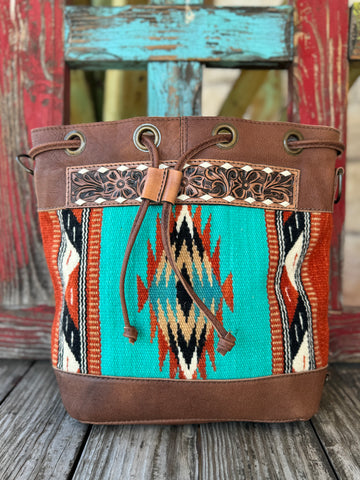 Women’s American Darling blanket leather bucket purse in turquoise and brown, with hand-tooled floral leather trim and woven wool panel, featuring adjustable strap and drawstring closure – ADBG862F. Available at Blair’s Western Wear, Marble Falls, TX.