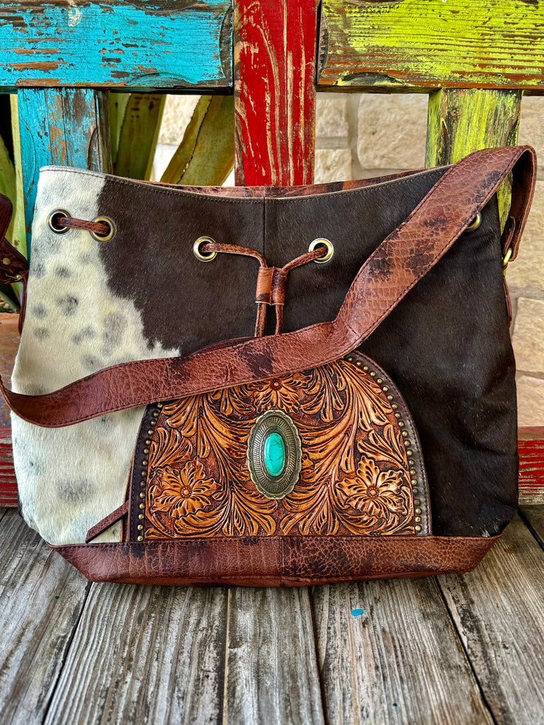 Women’s American Darling drawstring hide bag in brown and natural hair-on hide, featuring hand-tooled leather with a turquoise stone concho and a secure leather drawstring closure – ADBGI226B. Available at Blair's Western Wear, Marble Falls, TX.