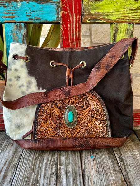 Women’s American Darling drawstring hide bag in brown and natural hair-on hide, featuring hand-tooled leather with a turquoise stone concho and a secure leather drawstring closure – ADBGI226B. Available at Blair's Western Wear, Marble Falls, TX.
