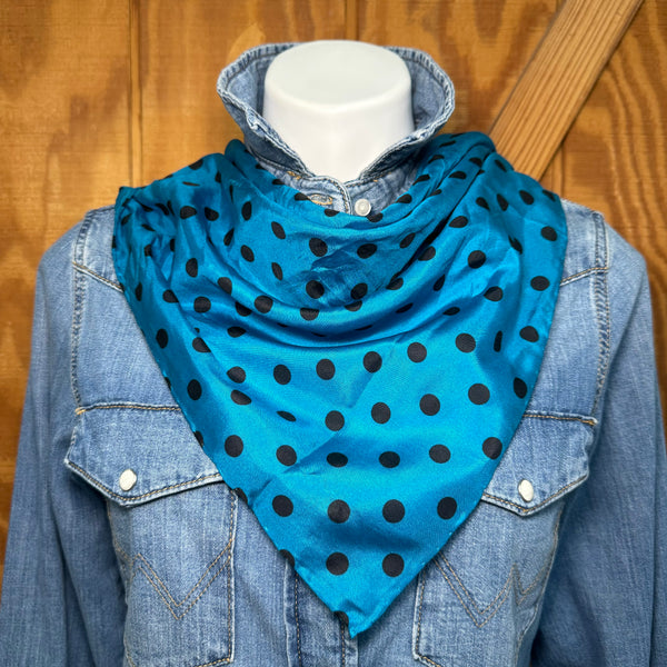 A bright blue silk-like bandana with black polka dots, styled on a white mannequin torso wearing a denim shirt against a wooden background.