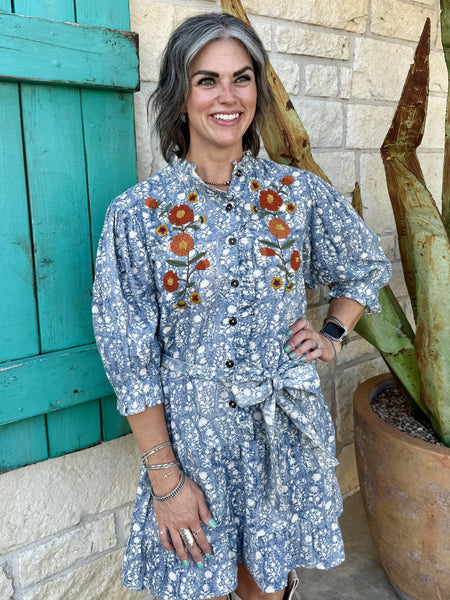 72703 — Women's blue-grey and white botanical block print tiered shirt dress with bold rust burnt orange and gold floral embroidery on bodice, black button-front placket, ruffle band collar, 3/4 puff sleeves with button cuffs, self-tie sash belt, and tiered ruffle hem skirt — Blair's Western Wear Marble Falls TX