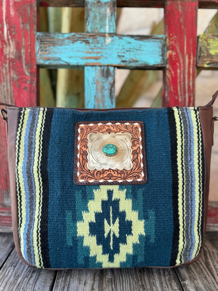 ADBGA286H — Women's messenger bag with a bold Navajo-inspired geometric wool front panel in deep teal, navy, lime green, and ivory, centered with a square hand-tooled leather frame, silver scalloped concho, and genuine turquoise stone — brown pebbled leather sides and adjustable tooled strap — Blair's Western Wear Marble Falls TX
