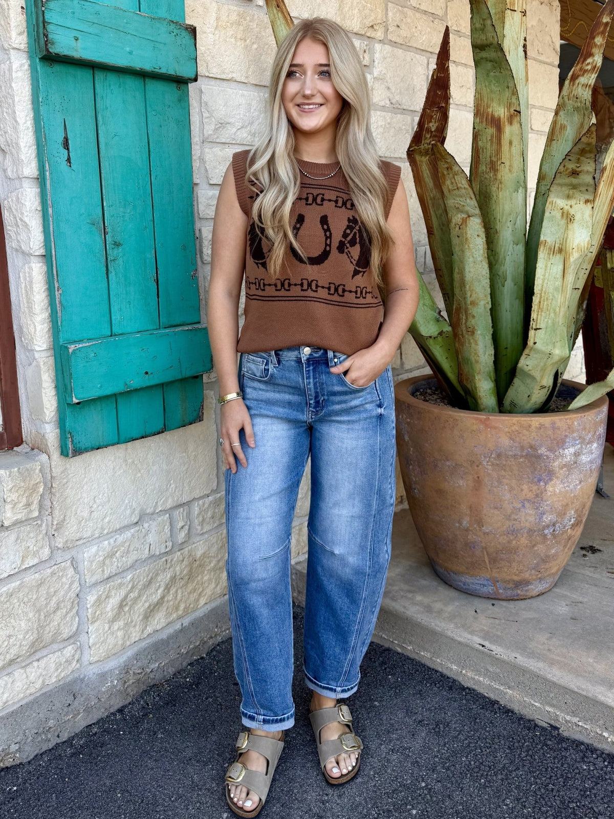 112382527 — Women's warm brown/caramel sleeveless knit sweater vest with bold intarsia knit western graphic featuring large center horseshoe flanked by horse heads and dark brown bit and chain-link border stripes, crew neckline with ribbed edge, structured shoulder seam, and ribbed hem band — Blair's Western Wear Marble Falls TX