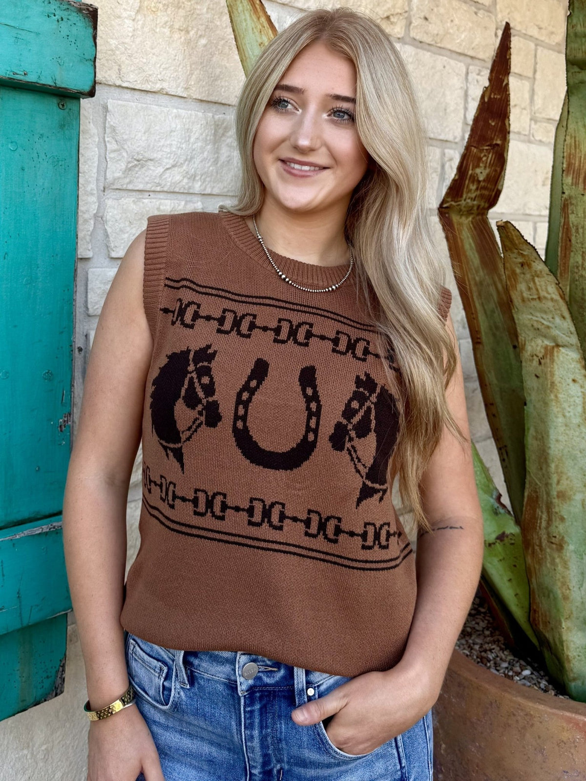 112382527 — Women's warm brown/caramel sleeveless knit sweater vest with bold intarsia knit western graphic featuring large center horseshoe flanked by horse heads and dark brown bit and chain-link border stripes, crew neckline with ribbed edge, structured shoulder seam, and ribbed hem band — Blair's Western Wear Marble Falls TX