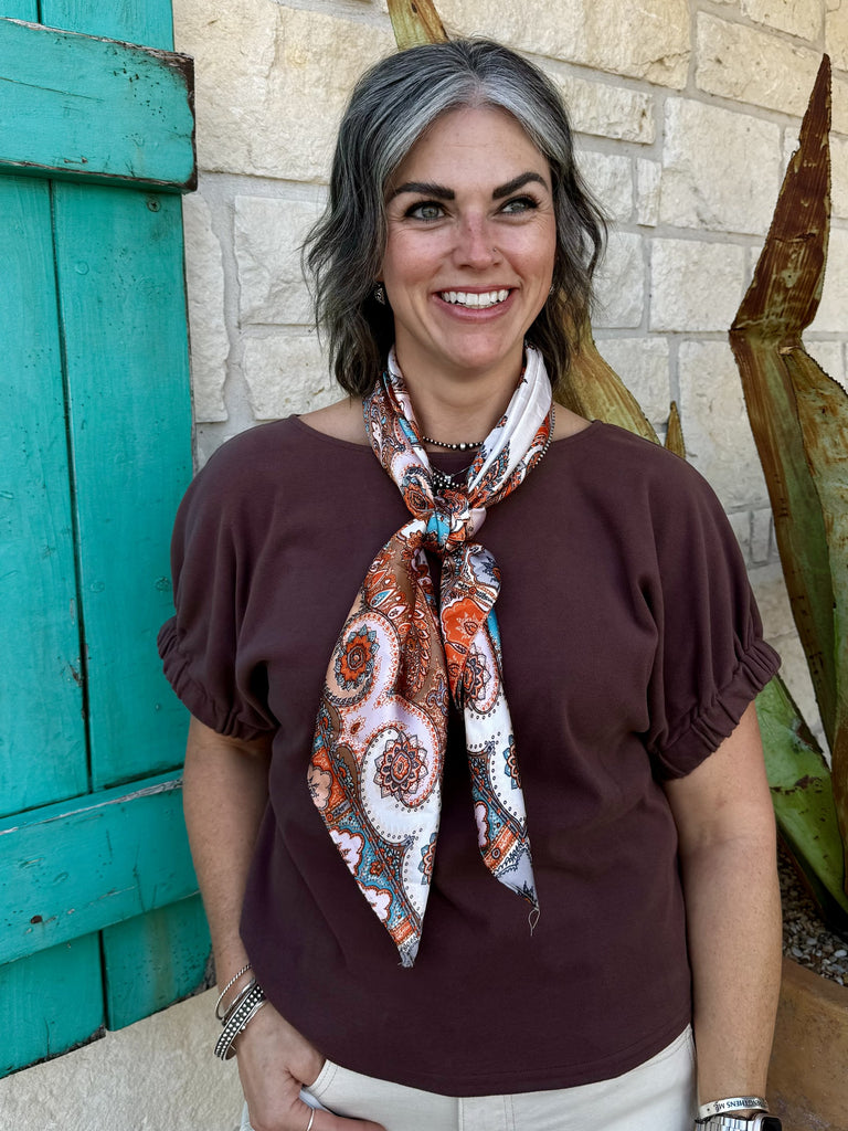 TK16456C — Women's chocolate brown soft knit crop top with wide scoop neckline, short puff sleeves with gathered elastic cuffs, and relaxed boxy cropped silhouette, styled with ivory wide leg jeans, paisley wild rag scarf, and silver Navajo pearl necklace — Blair's Western Wear Marble Falls TX