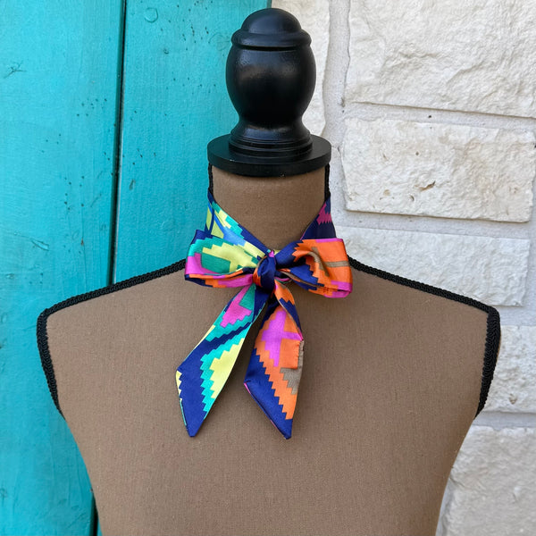 A multicolor geometric print neck scarf tied around a brown dress form mannequin, set against a background of turquoise painted wood and a white stone wall.
