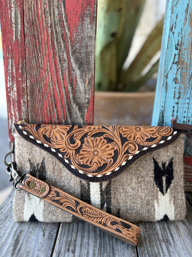 ADBG1477C — Front view of women's gray Navajo-inspired blanket wool envelope wristlet clutch showing heathered gray/taupe wool body with black and cream geometric motifs, pale natural tan hand-tooled leather envelope flap with floral and scroll tooling on black background, dark chocolate leather border trim with cream whipstitch edge, and tooled leather wrist strap with antique silver snap hardware — Blair's Western Wear Marble Falls TX