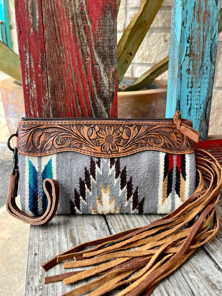 ADBG344D4 — Women's grey Aztec-inspired blanket wool wristlet with bold feather and arrow geometric pattern in turquoise/teal, navy, white, black, chocolate brown, red, and tan/gold, wide medium brown hand-tooled leather top yoke featuring central sunflower medallion with sweeping acanthus scroll and leaf tooling and scalloped bottom edge, long tan/caramel suede leather fringe on the right side, gunmetal zipper with leather pull tab, and gunmetal swivel clip wristlet strap on the left — Blair's Western Wear