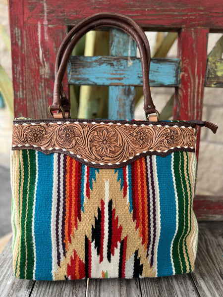 ADBGA151G — Women's large structured tote with bold multicolor Aztec-inspired serape woven fabric body in turquoise/aqua, green, orange, red, cream, gold, black, and purple with a large central geometric arrow and chevron medallion, wide pale natural tan hand-tooled leather top yoke featuring central daisy/sunflower medallion with floral and acanthus scroll tooling and dark chocolate leather edge trim with cream whipstitch detail, and dark brown rolled leather top handles with antique gold D-ring hardware —
