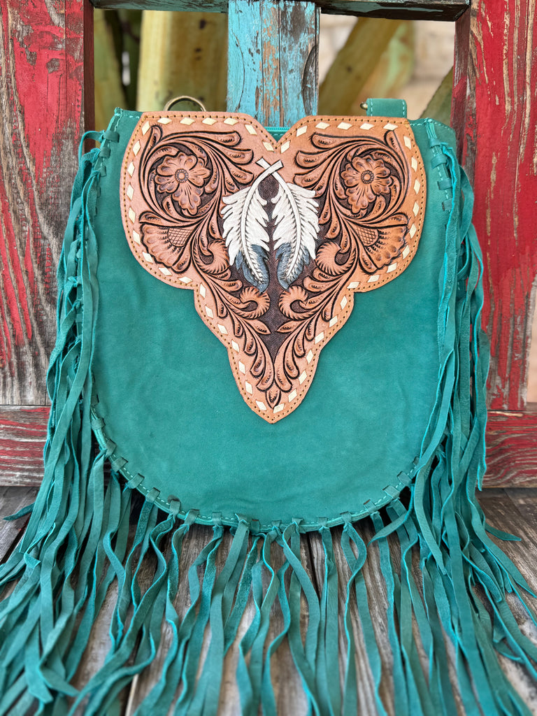 ADBGA431E — This jaw-dropping turquoise suede crossbody has it all — long turquoise fringe on both sides and bottom, a sculpted tan hand-tooled floral leather flap with a painted black and white feather centerpiece, and gold lace whipstitch trim that makes it pure boho western art — Blair's Western Wear Marble Falls TX