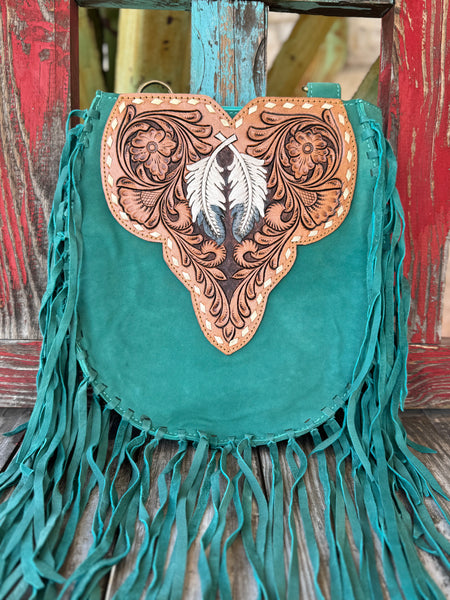 ADBGA431E — This jaw-dropping turquoise suede crossbody has it all — long turquoise fringe on both sides and bottom, a sculpted tan hand-tooled floral leather flap with a painted black and white feather centerpiece, and gold lace whipstitch trim that makes it pure boho western art — Blair's Western Wear Marble Falls TX