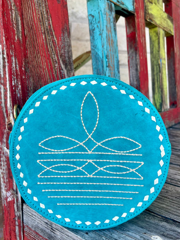 ADBGA620A — Women's round turquoise/teal suede crossbody bag with intricate white western boot toe stitch pattern on the front featuring a central leaf-shaped medallion, three rows of interlocking elongated oval motifs, and horizontal running stitch lines, white diamond whipstitch lacing running the full circumference edge, and perimeter zipper closure — Blair's Western Wear Marble Falls TX