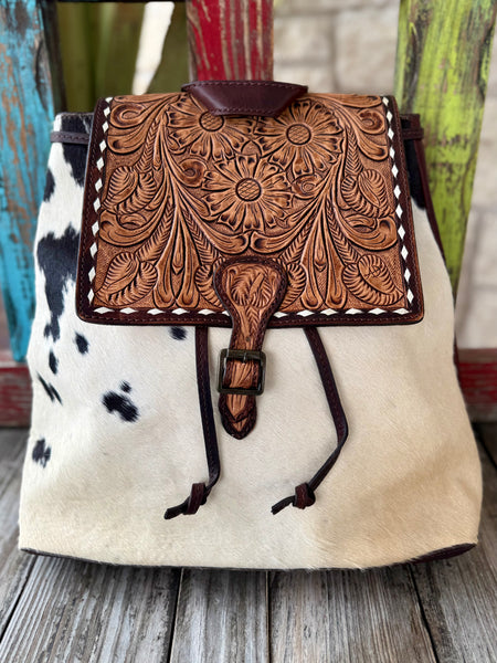 ADBGZ102TAW — Women's structured western backpack with crisp white and black spotted hair-on cowhide body, full-coverage hand-tooled tan leather flap featuring three large sunflower/daisy medallions with sweeping acanthus scroll, feather, and leaf carving, dark chocolate/burgundy brown leather whipstitch lacing on all four flap edges, tooled leather tongue strap with gunmetal buckle and knotted leather drawstrings, small leather loop handle at top center, and adjustable dark brown leather shoulder straps — 