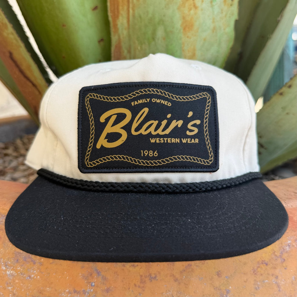 A black and off- white snapback cap with a prominent black front patch featuring gold embroidered text that reads 'Blair's Western Wear 1986'.
