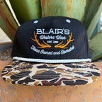A black snapback cap with 'BLAIR'S Western Wear' embroidered on the front, featuring orange antler graphics and a brown, beige, and black camouflage patterned brim, set against a background of green succulent leaves.