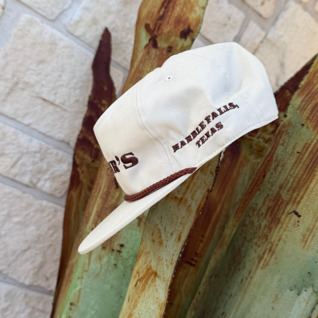 An off-white baseball cap with brown embroidered text reading 'MARBLE FALLS, TEXAS' and a matching brown rope detail on the brim