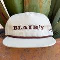 An off-white snapback cap with 'BLAIR'S' embroidered in dark brown across the front, featuring a matching brown braided rope detail around the crown and a flat brim