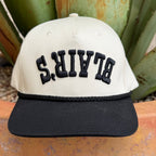 A structured baseball cap with an off-white crown and a black flat brim, featuring the word 'BLAIR'S' embroidered in black arched letters across the front updside down 