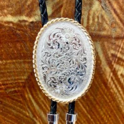 Men's Western bolo tie with silver and gold scroll design, featuring an engraved silver oval concho with gold rope edges, black braided leather cord, and polished silver tips - 22267. Available at Blair's Western Wear, Marble Falls, TX.

