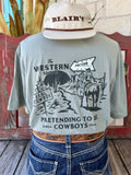 Men's sage green Sendero Provisions Co. The Western Show short sleeve tee with black cowboy on horseback crossroads chest pocket graphic and Pretending to Be Cowboys Since 2014 text, front view styled with jeans and tooled leather belt — Blair's Western Wear Marble Falls TX