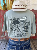Men's sage green Sendero Provisions Co. The Western Show short sleeve tee with black cowboy on horseback crossroads chest pocket graphic and Pretending to Be Cowboys Since 2014 text, front view styled with jeans and tooled leather belt — Blair's Western Wear Marble Falls TX