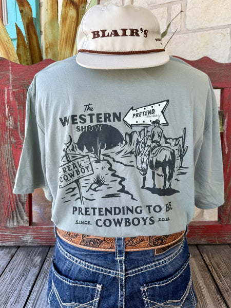 Men's sage green Sendero Provisions Co. The Western Show short sleeve tee with black cowboy on horseback crossroads chest pocket graphic and Pretending to Be Cowboys Since 2014 text, front view styled with jeans and tooled leather belt — Blair's Western Wear Marble Falls TX