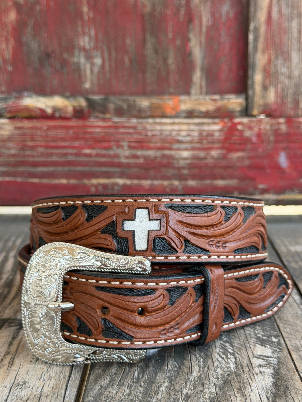 Two-toned brown and black tooled leather features sweeping floral scrollwork and a standout white hair-on-hide cross inlay, finished with cream stitching and an engraved silver buckle — bold western faith and craftsmanship in one. Available at BLAIR'S Western Wear located in Marble Falls TX 