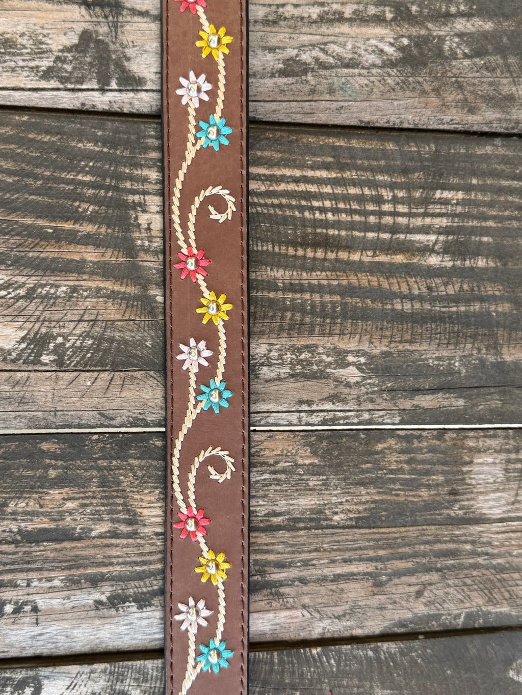 A brown leather belt with white embroidered swirls and colorful floral details, featuring a large engraved silver buckle, displayed on a rustic wooden surface. Available at BLAIR'S Western Wear located in Marble Falls TX 