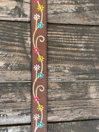A brown leather belt with white embroidered swirls and colorful floral details, featuring a large engraved silver buckle, displayed on a rustic wooden surface. Available at BLAIR'S Western Wear located in Marble Falls TX 