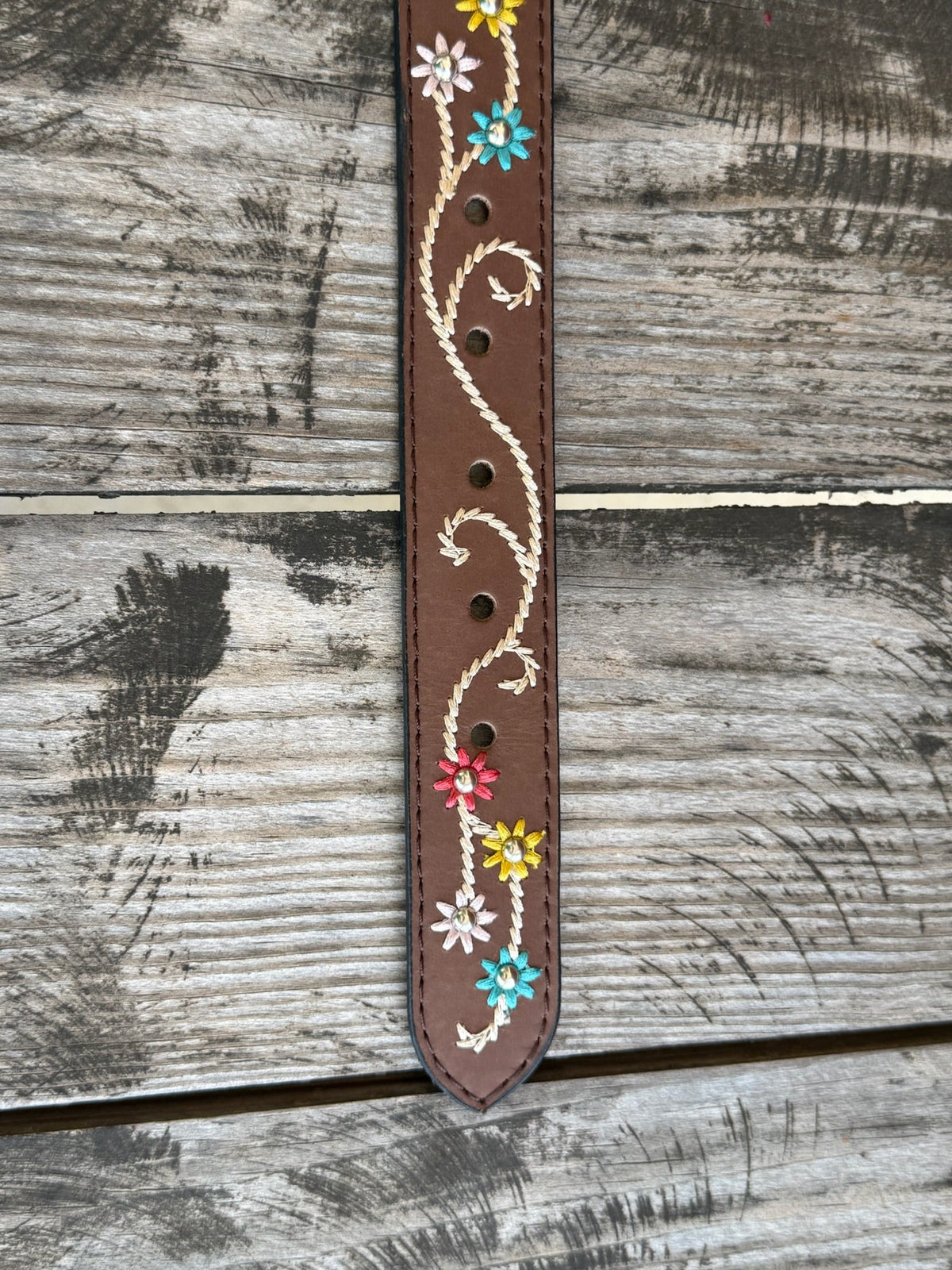 A brown leather belt with white embroidered swirls and colorful floral details, featuring a large engraved silver buckle, displayed on a rustic wooden surface. Available at BLAIR'S Western Wear located in Marble Falls TX 