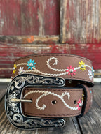 A brown leather belt with white embroidered swirls and colorful floral details, featuring a large engraved silver buckle, displayed on a rustic wooden surface. Available at BLAIR'S Western Wear located in Marble Falls TX 
