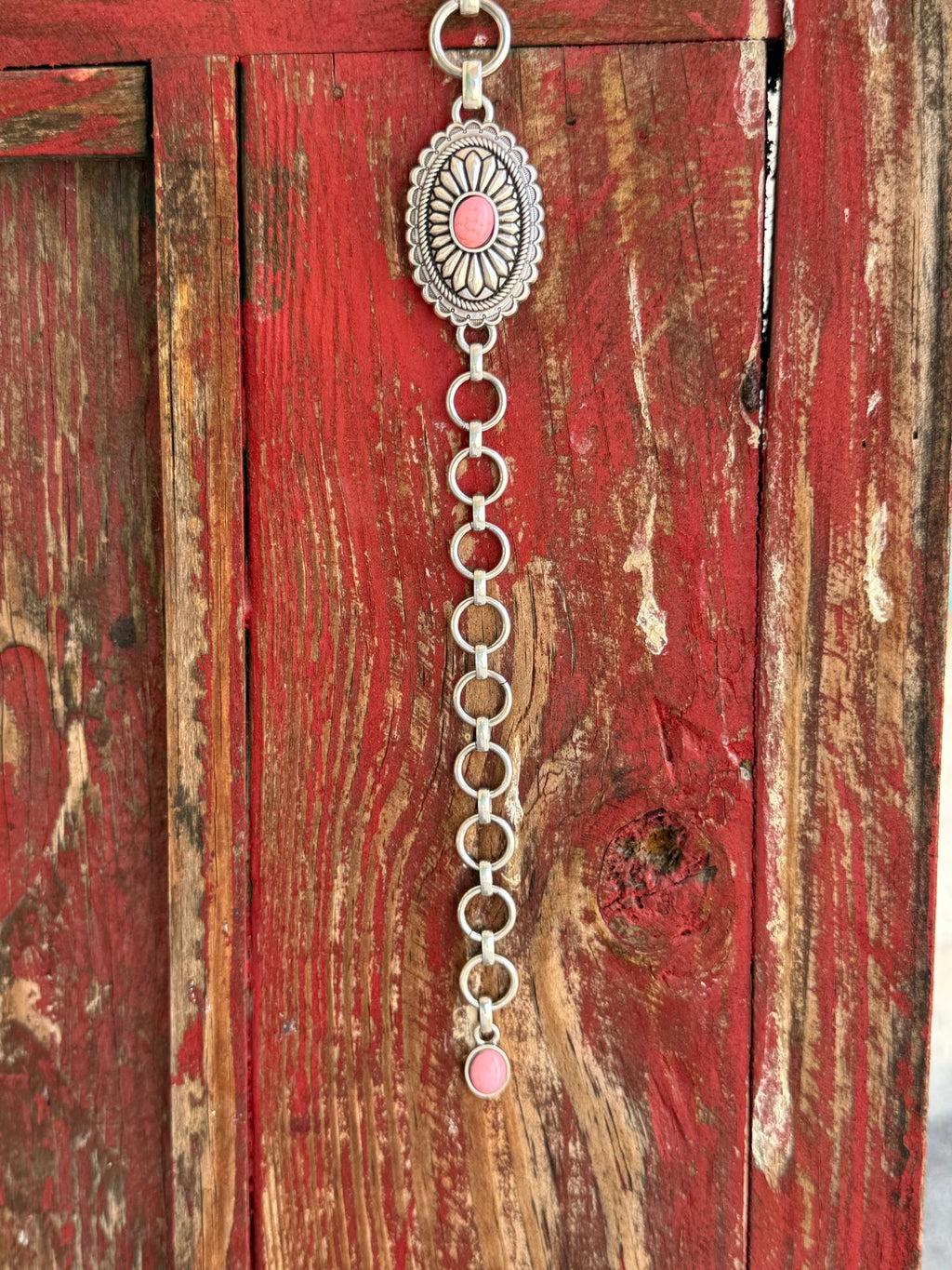 A1308230 — Kid's Pink Concho Chain Western Belt Full silver concho chain belt with repeating oval silver conchos featuring engraved petal/sunburst detail, rope edge border, and pink stone center in each. Silver O-ring links connect each concho. Adjustable silver O-ring chain tail with small pink stone drop tip. Available at Blair's Western Wear located in Marble Falls TX 