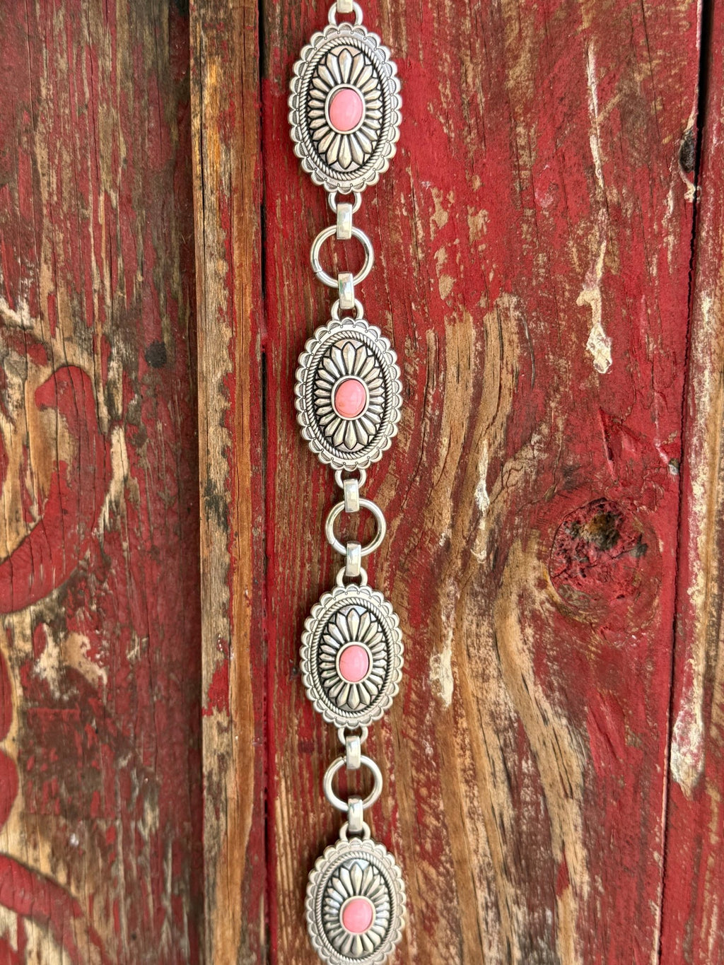 A1308230 — Kid's Pink Concho Chain Western Belt Full silver concho chain belt with repeating oval silver conchos featuring engraved petal/sunburst detail, rope edge border, and pink stone center in each. Silver O-ring links connect each concho. Adjustable silver O-ring chain tail with small pink stone drop tip. Available at Blair's Western Wear located in Marble Falls TX 