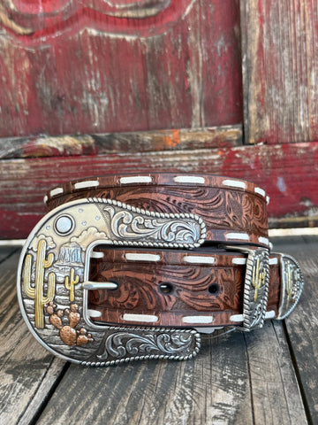 A western-style leather belt with a carved brown leather body, silver-toned engraved buckle, and white contrast stitching, displayed on a rustic wooden surface in front of a weathered red wooden door.