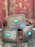A western-style belt with large engraved silver-tone square buckles, each featuring a round turquoise stone, resting on a rustic wooden surface with a blurred red decorative background.
